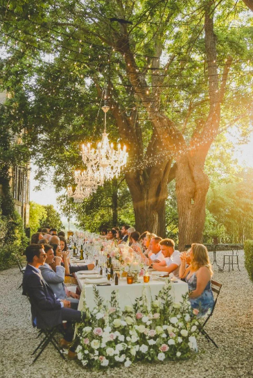 Lange feesttafel onder grote bomen met kroonluchters en lichtjes